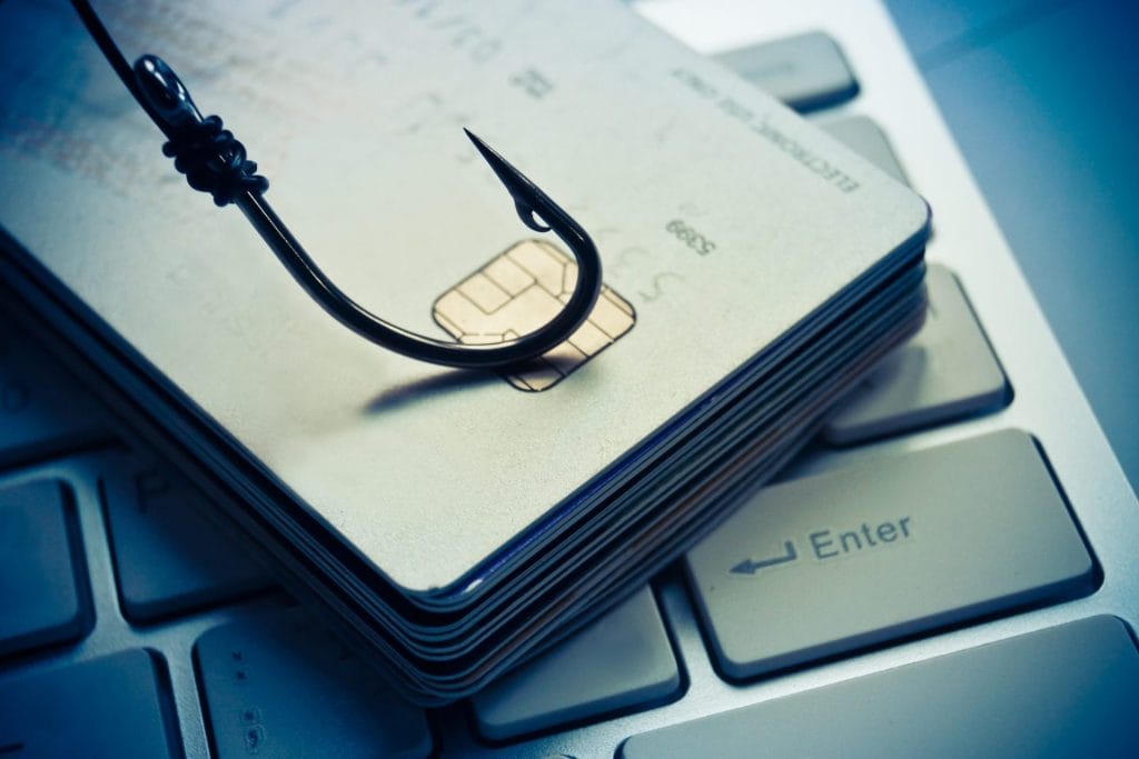 Gomila kreditnih kartica postavljenih na tastaturu laptopa, sa ribarskom udicom zakačenom za njih. Ovo simbolizuje phishing napad, koji je uobičajena prevara na sajber sigurnost gde napadači pokušavaju da prevare ljude kako bi otkrili njihove lične informacije.