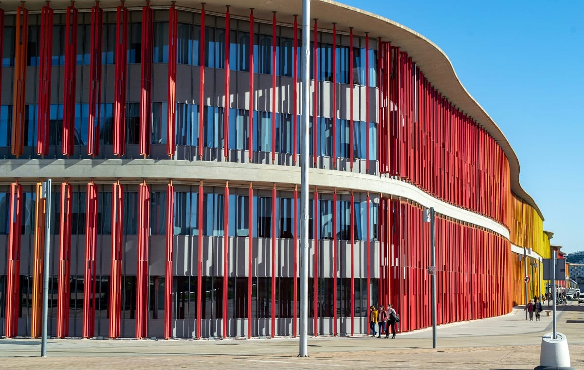 A modern, colorful building with curved architecture, featuring vertical red, orange, and yellow slats along its glass facade. People walk along the pavement in front of the building under a clear blue sky.
