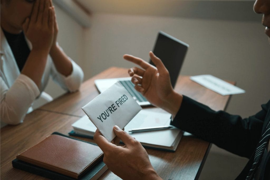Poslodavac uručuje kovertu s natpisom "you're fired" zaposlenom koji deluje iznenađeno, što simbolizuje otkazni rok. Na stolu su laptop, rokovnik i poslovna dokumentacija.
