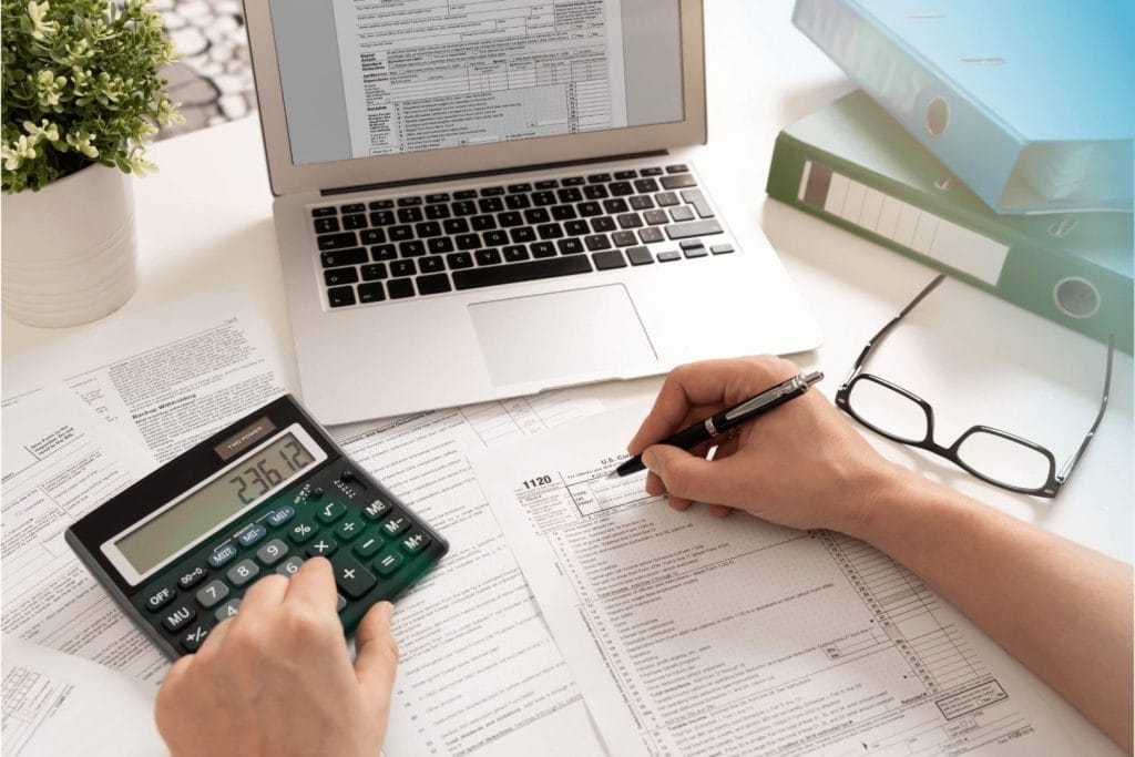 Osoba koristi digitron i popunjava poresku prijavu. Na stolu se nalaze laptop, naočare, fascikle i biljka. Ilustracija simbolizuje prihode pomoraca i oporezivanje pomoraca.