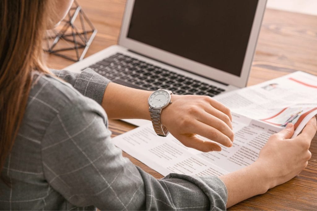 Ženska gleda na uro, medtem ko drži časopis, sedi ob prenosniku na mizi