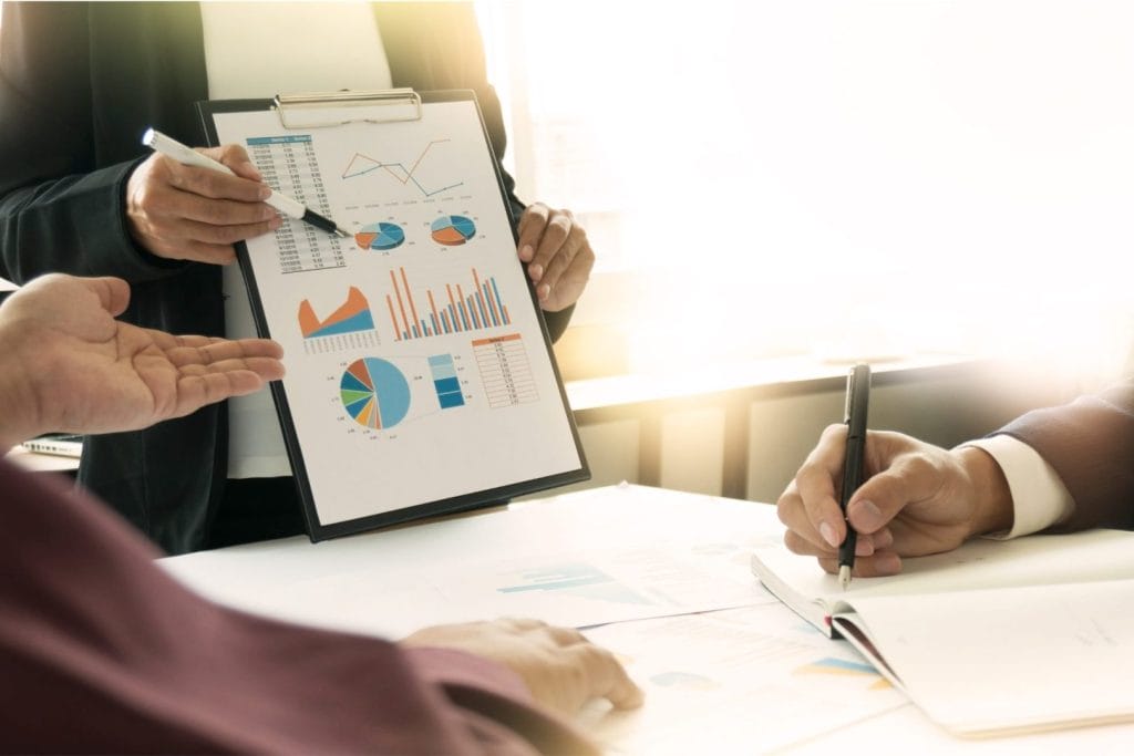 Biznismeni u diskusiji analiziraju finansijske izveštaje i grafikone. Jedna osoba drži tablu sa poslovnim podacima, dok druga beleži napomene, simbolizujući strateško planiranje i investicione odluke.