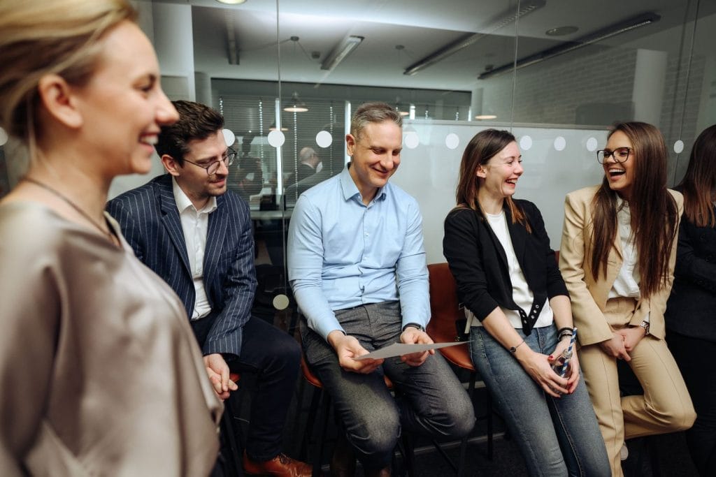 Grupa ljudi sedi i razgovara u kancelariji, smešeći se i uživajući u prijatnoj atmosferi.