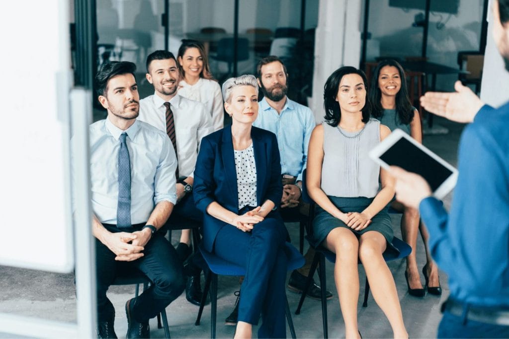 Grupa ljudi u formalnoj odeći sedi i sluša predavanje u modernoj kancelariji. Ilustracija simbolizuje korišćenje subvencija za usavršavanje i zapošljavanje.