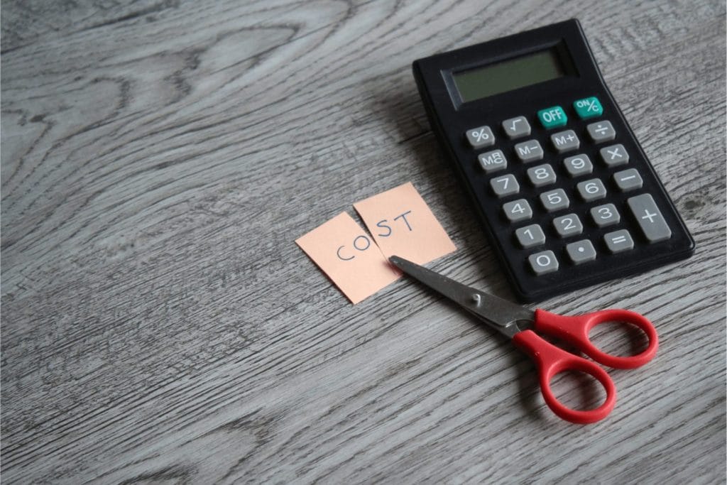 Makaze sijeku papir s natpisom 'COST', pored digitrona na drvenoj podlozi.