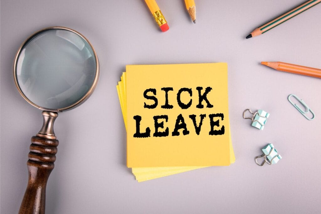 Žuti post-it sa natpisom "SICK LEAVE" (bolovanje), postavljen na radnoj površini, okružen raznim kancelarijskim priborom poput lupa, olovaka, spajalica i dugmadi za papir.